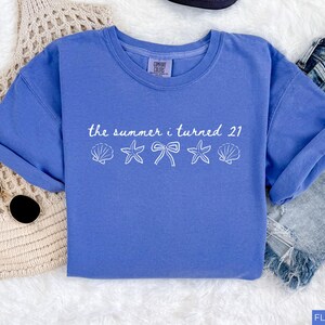 Pu&ograve; includere: T-shirt blu con la scritta "the summer i turned 21" e grafiche di conchiglie, stelle marine e fiocchi. Le maniche sono arrotolate. La maglietta &egrave; accanto a un paio di occhiali da sole e pantaloncini di jeans.