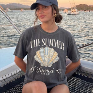 May include: A charcoal gray short-sleeve t-shirt featuring a large central graphic of a seashell with blue, yellow, and white floral and plaid patterns. The text reads THE SUMMER I Turned 13 EST 2013 in white lettering above and below the shell.