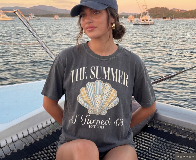 May include: A charcoal gray short-sleeve t-shirt featuring a large central graphic of a seashell with blue, yellow, and white floral and plaid patterns. The text reads THE SUMMER I Turned 13 EST 2013 in white lettering above and below the shell.
