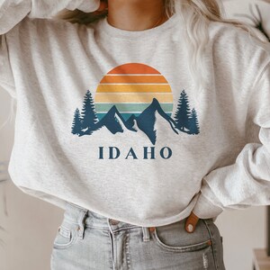 May include: A gray sweatshirt with a retro sunset graphic and mountains. The text "IDAHO" is printed below the graphic.