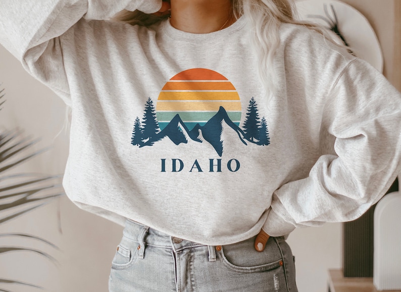May include: A gray sweatshirt with a retro sunset graphic and mountains. The text "IDAHO" is printed below the graphic.