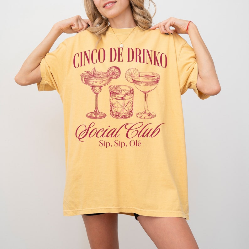 May include: A yellow t-shirt with a red graphic print that says 'Cinco De Drinko Social Club Sip, Sip, Ole' with a drawing of three cocktails.