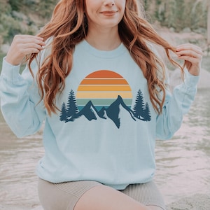May include: A light blue sweatshirt with a retro sunset and mountain graphic. The graphic features a multi-colored sunset with a silhouette of mountains and trees.