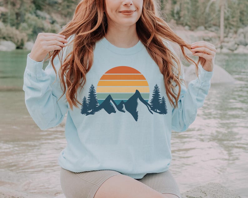 May include: A light blue sweatshirt with a retro sunset and mountain graphic. The graphic features a multi-colored sunset with a silhouette of mountains and trees.