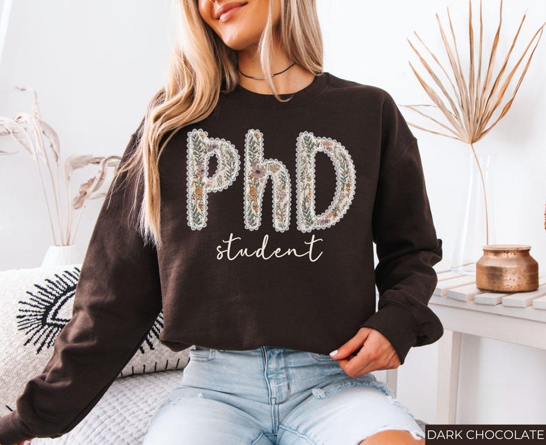 May include: A dark chocolate brown sweatshirt featuring the text "PhD student". The letters "PhD" are decorated with a floral design, and the word "student" is written in a delicate script font.