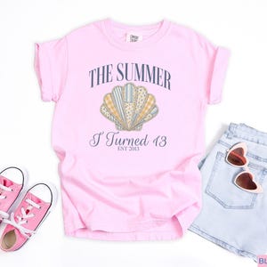 May include: A light pink short-sleeve t-shirt featuring a graphic of a seashell with various patterned segments. The text reads THE SUMMER I Turned 13 EST 2013. The shirt is displayed with light blue denim shorts, pink canvas sneakers, and heart-shaped sunglasses.