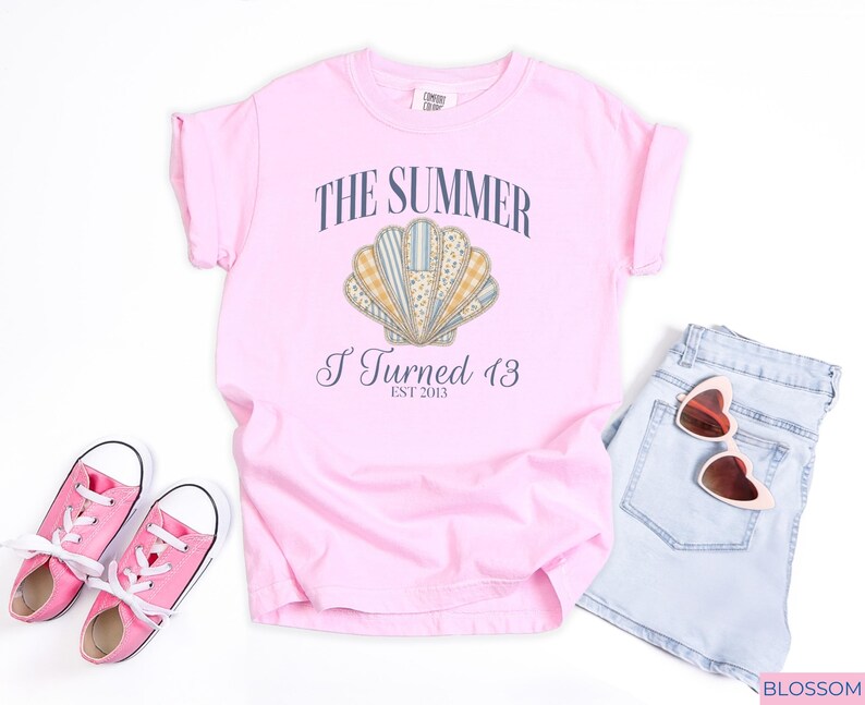 May include: A light pink short-sleeve t-shirt featuring a graphic of a seashell with various patterned segments. The text reads THE SUMMER I Turned 13 EST 2013. The shirt is displayed with light blue denim shorts, pink canvas sneakers, and heart-shaped sunglasses.