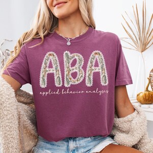 May include: A maroon t-shirt with the letters "ABA" in a floral design. Below the letters, the words "applied behavior analysis" are written in a script font. The shirt is a casual style.