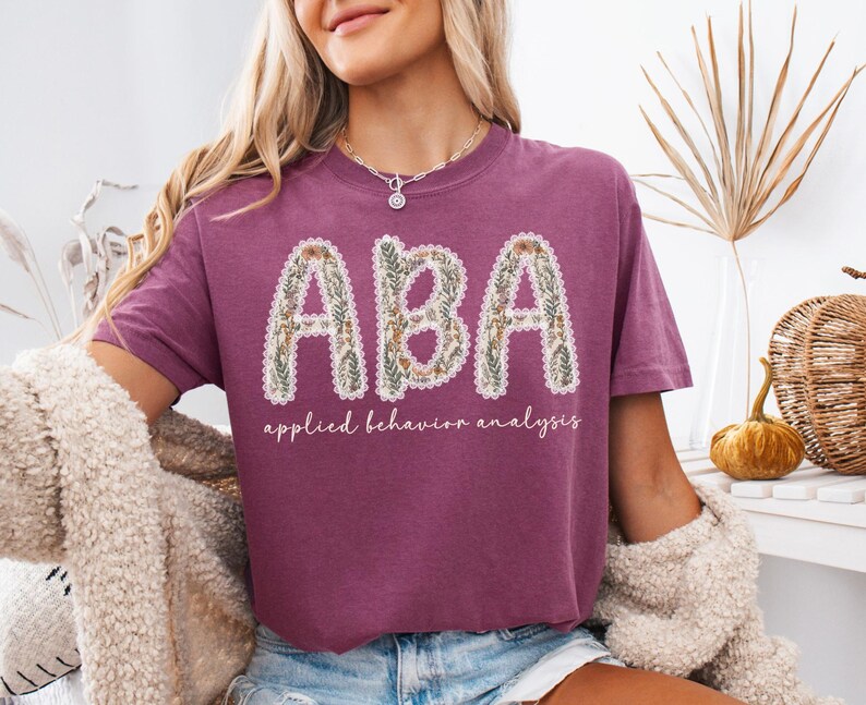 May include: A maroon t-shirt with the letters "ABA" in a floral design. Below the letters, the words "applied behavior analysis" are written in a script font. The shirt is a casual style.