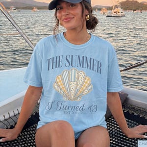 May include: A light blue short-sleeve t-shirt featuring a graphic of a seashell composed of various patterned segments in yellow, white, and blue. The text reads THE SUMMER I TURNED 13 EST 2013 in a dark blue font centered above and below the seashell graphic.