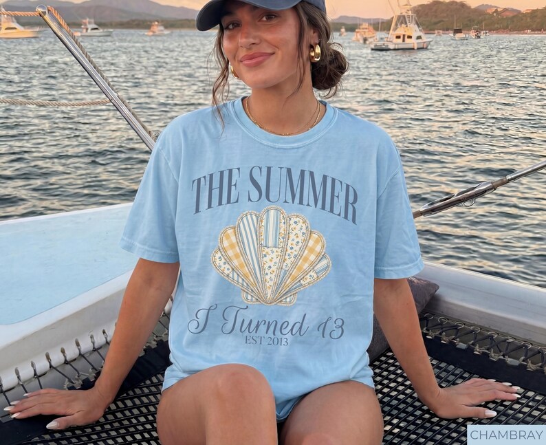 May include: A light blue short-sleeve t-shirt featuring a graphic of a seashell composed of various patterned segments in yellow, white, and blue. The text reads THE SUMMER I TURNED 13 EST 2013 in a dark blue font centered above and below the seashell graphic.
