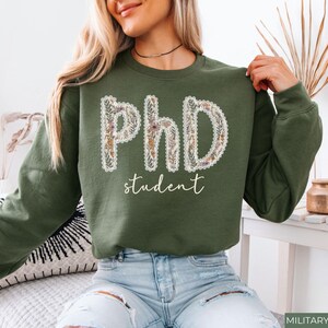 May include: An olive green sweatshirt featuring the words "PhD student". The letters are decorated with a floral pattern and a white lace trim. The word "student" is written in a cursive font. The sweatshirt is a casual garment.