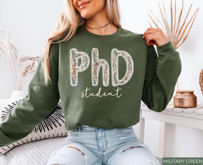 May include: An olive green sweatshirt featuring the words "PhD student". The letters are decorated with a floral pattern and a white lace trim. The word "student" is written in a cursive font. The sweatshirt is a casual garment.