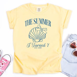 K&ouml;nnte beinhalten: Ein hellgelbes T-Shirt mit dem Text "THE SUMMER I TURNED 7 EST 2019" und einer Muschel-Grafik. Das Bild zeigt rosa Turnschuhe, Jeansshorts und herzf&ouml;rmige Sonnenbrillen.