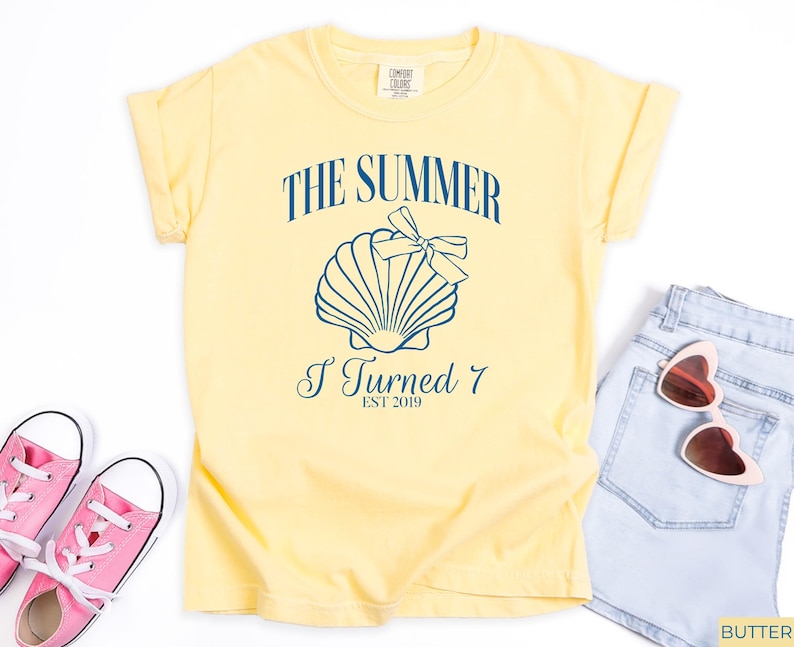 K&ouml;nnte beinhalten: Ein hellgelbes T-Shirt mit dem Text "THE SUMMER I TURNED 7 EST 2019" und einer Muschel-Grafik. Das Bild zeigt rosa Turnschuhe, Jeansshorts und herzf&ouml;rmige Sonnenbrillen.