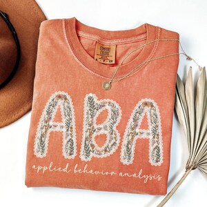 May include: Coral-colored t-shirt with the letters "ABA" in a floral design, and the words "applied behavior analysis" below. A gold necklace and a brown hat are also in the image.