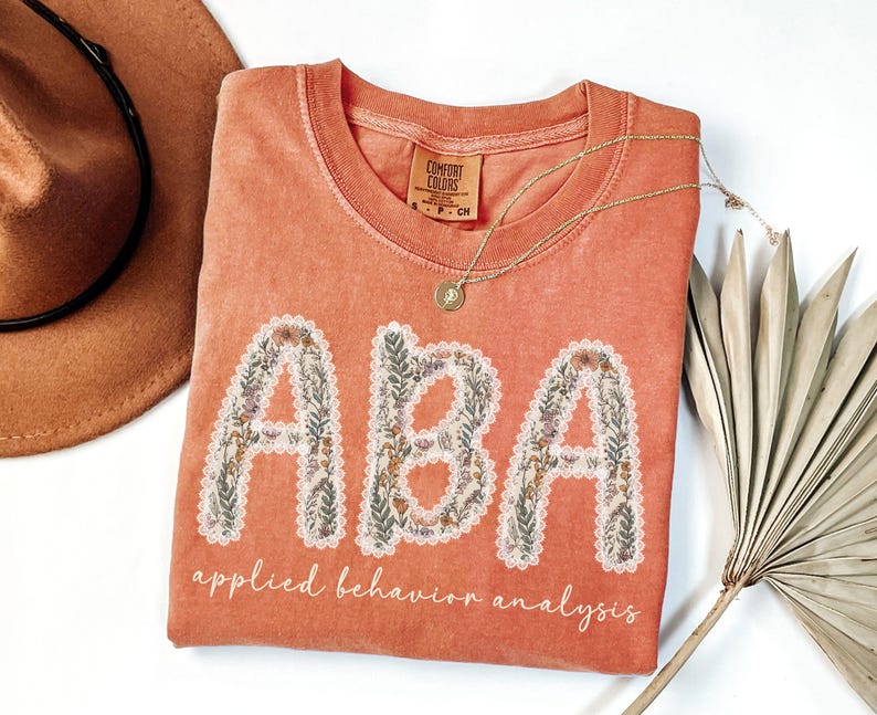 May include: Coral-colored t-shirt with the letters "ABA" in a floral design, and the words "applied behavior analysis" below. A gold necklace and a brown hat are also in the image.