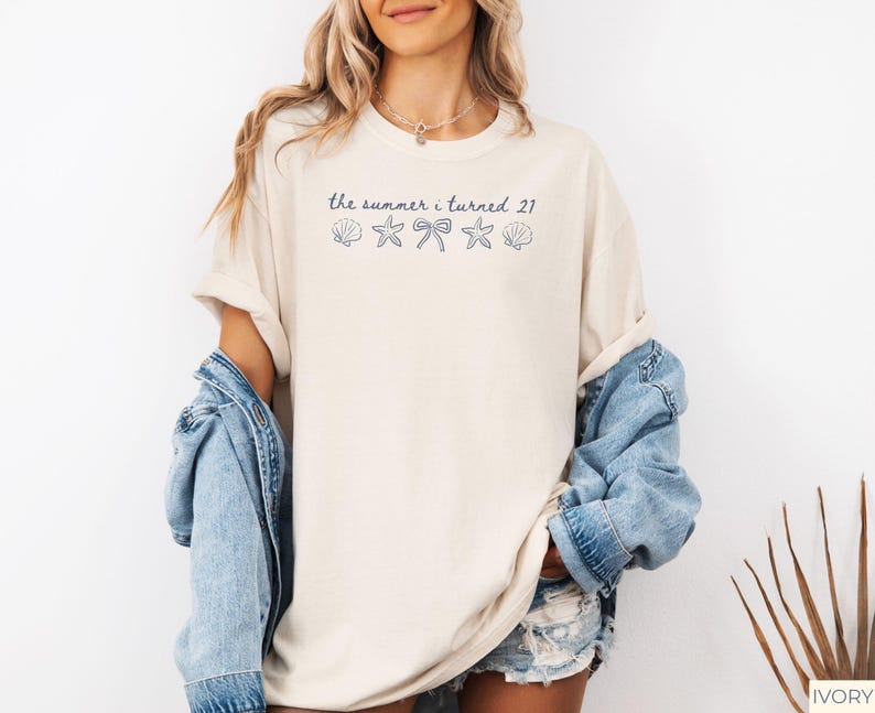 Pu&ograve; includere: T-shirt color crema con la scritta blu navy "the summer i turned 21" e grafiche di conchiglie, fiocco e stella marina. La maglietta &egrave; indossata con una giacca di jeans.