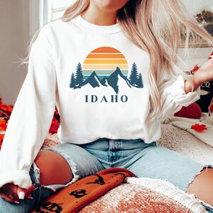 May include: White sweatshirt with a graphic design of mountains, a sunset, and the word "IDAHO".