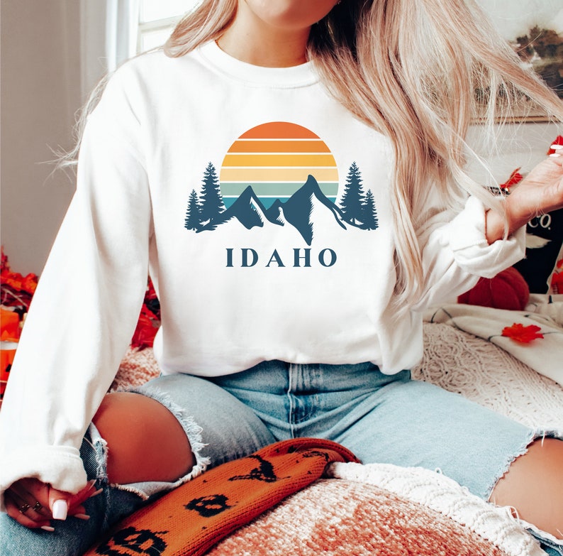 May include: White sweatshirt with a graphic design of mountains, a sunset, and the word "IDAHO".