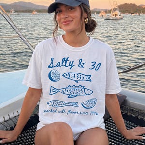 May include: White t-shirt with blue fish and lemon slice graphics, and the text "Salty & 30" and "packed with flavour since 1996". The person is wearing a blue cap and gold earrings.