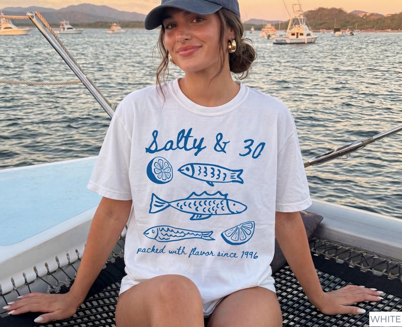 May include: White t-shirt with blue fish and lemon slice graphics, and the text "Salty & 30" and "packed with flavour since 1996". The person is wearing a blue cap and gold earrings.