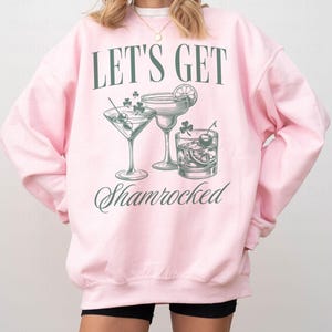以下が含まれることがあります： 「Let's Get Shamrocked」という文字と、3つのカクテルグラスのグラフィックが描かれたピンク色のスウェットシャツ。