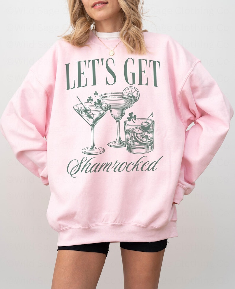 以下が含まれることがあります： 「Let's Get Shamrocked」という文字と、3つのカクテルグラスのグラフィックが描かれたピンク色のスウェットシャツ。