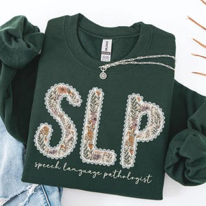 May include: A dark green sweatshirt with the letters "SLP" in a floral design. The words "speech language pathologist" are printed below the letters. A silver necklace with a pendant is draped across the sweatshirt.