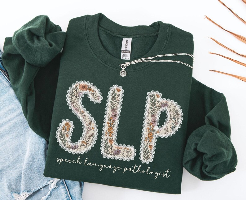 May include: A dark green sweatshirt with the letters "SLP" in a floral design. The words "speech language pathologist" are printed below the letters. A silver necklace with a pendant is draped across the sweatshirt.