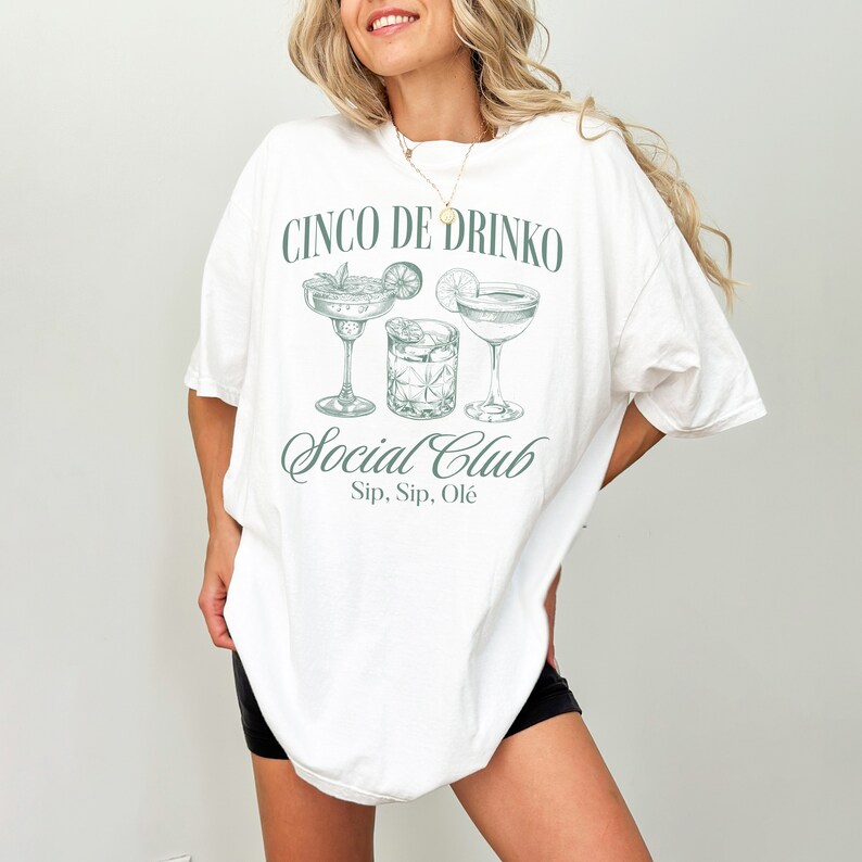 May include: White oversized t-shirt with a green graphic of three cocktail glasses and the text "Cinco de Drinko Social Club Sip, Sip, Ol&egrave;"