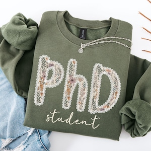 May include: An olive green sweatshirt featuring the text "PHD student" in a floral design. The letters are filled with flowers in yellow, orange, and purple. A silver necklace with a small pendant is draped across the sweatshirt.