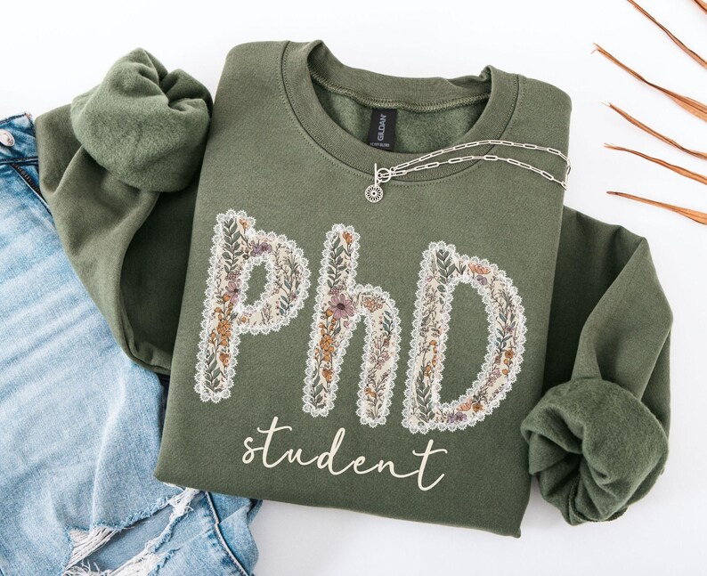 May include: An olive green sweatshirt featuring the text "PHD student" in a floral design. The letters are filled with flowers in yellow, orange, and purple. A silver necklace with a small pendant is draped across the sweatshirt.