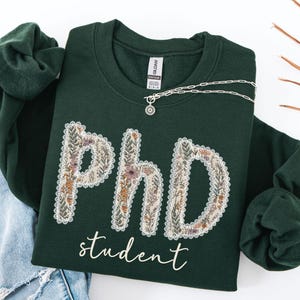 May include: A dark green sweatshirt featuring the text "PhD student" in white script. The letters "PhD" are decorated with a floral pattern in cream, brown, and purple. A silver necklace with a pendant is draped across the sweatshirt.