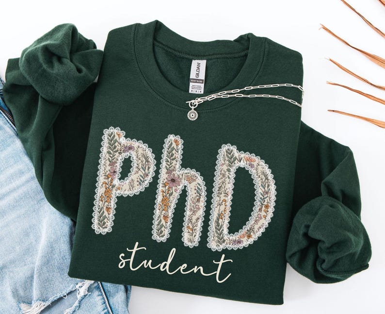 May include: A dark green sweatshirt featuring the text "PhD student" in white script. The letters "PhD" are decorated with a floral pattern in cream, brown, and purple. A silver necklace with a pendant is draped across the sweatshirt.