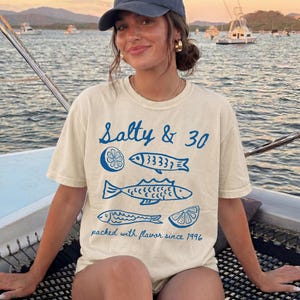 May include: Ivory t-shirt featuring the text "Salty & 30" and blue fish and lemon slice illustrations. The phrase "packed with flavour since 1996" is printed below. The shirt is worn on a boat, with a seascape in the background.