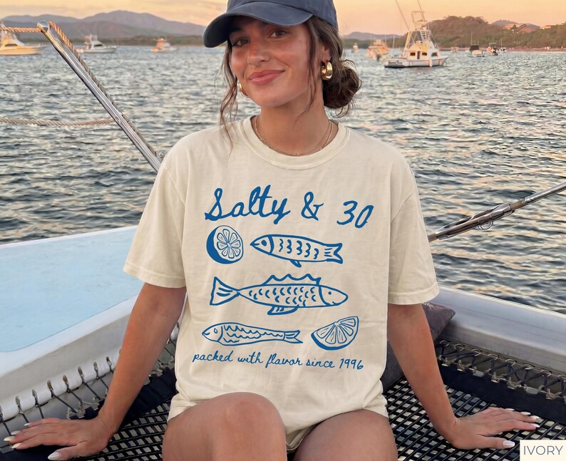 May include: Ivory t-shirt featuring the text "Salty & 30" and blue fish and lemon slice illustrations. The phrase "packed with flavour since 1996" is printed below. The shirt is worn on a boat, with a seascape in the background.
