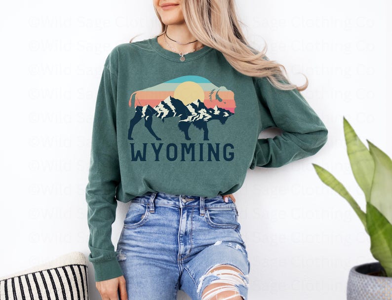 May include: A green long-sleeved shirt with a graphic of a bison in front of a mountain range with a sunset behind it. The text "WYOMING" is printed below the graphic.