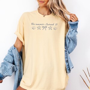 Pu&ograve; includere: T-shirt giallo chiaro con la scritta "the summer i turned 21" e grafiche di conchiglie, fiocchi e stelle marine. La maglietta &egrave; abbinata a una giacca di jeans azzurra.