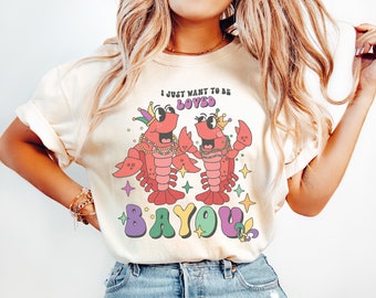 May include: A white t-shirt with a cartoon crawfish wearing a crown and beads. The crawfish is holding a sign that says "I just want to be loved." Below the crawfish is the word "BAYOU" in colorful letters.