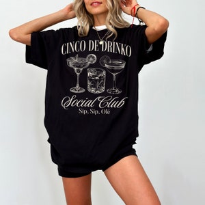 May include: Black oversized t-shirt with a graphic of three cocktail glasses and the text "Cinco de Drinko Social Club Sip, Sip, Ol&egrave;"