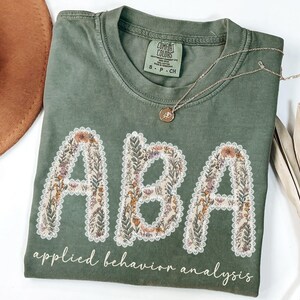 May include: Olive green t-shirt with the letters "ABA" in a floral design, outlined with white lace. The words "applied behavior analysis" are printed below the letters in a cursive font. A gold necklace rests on the shirt.