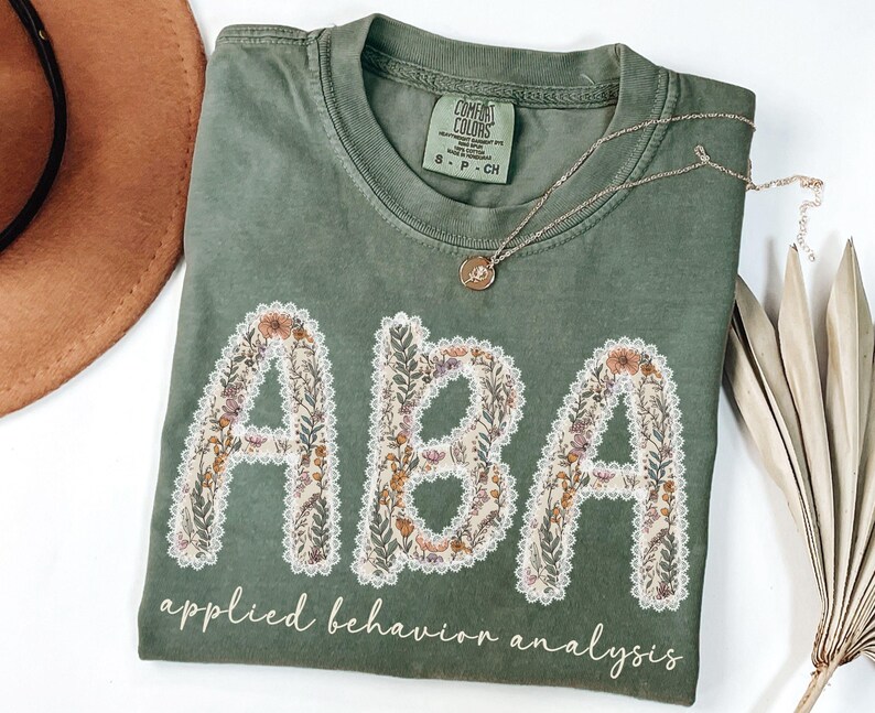 May include: Olive green t-shirt with the letters "ABA" in a floral design, outlined with white lace. The words "applied behavior analysis" are printed below the letters in a cursive font. A gold necklace rests on the shirt.