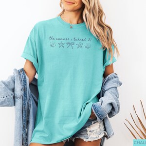 Pu&ograve; includere: Una maglietta turchese con le maniche arrotolate. La maglietta presenta la scritta "the summer i turned 21" sopra grafiche di conchiglie, fiocchi e stelle marine. La modella indossa una giacca di jeans e pantaloncini.