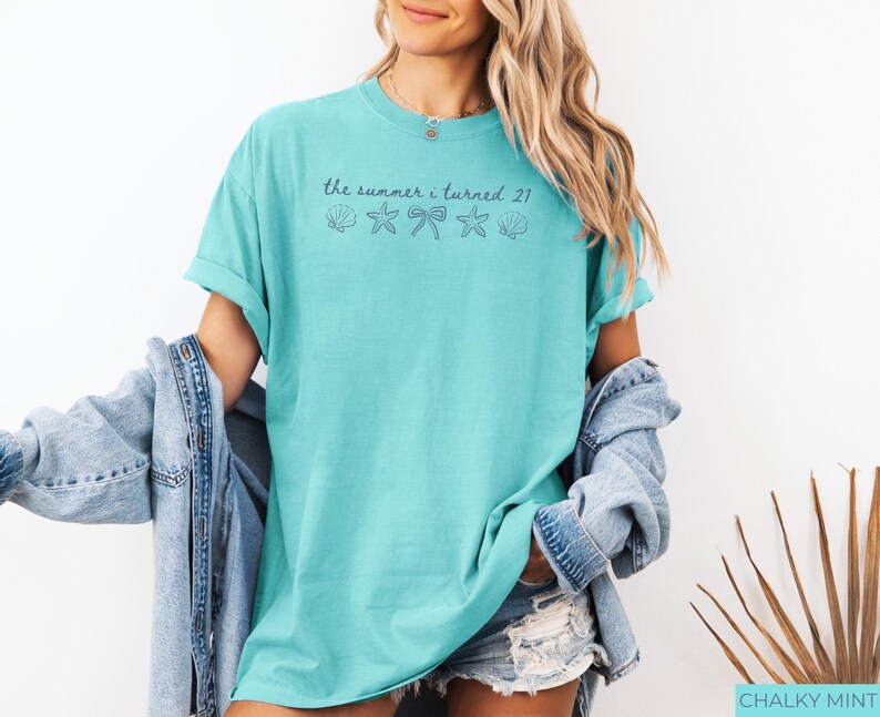 Pu&ograve; includere: Una maglietta turchese con le maniche arrotolate. La maglietta presenta la scritta "the summer i turned 21" sopra grafiche di conchiglie, fiocchi e stelle marine. La modella indossa una giacca di jeans e pantaloncini.