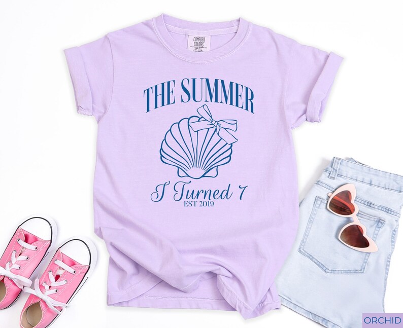 K&ouml;nnte beinhalten: Helllila T-Shirt mit dem Text "THE SUMMER I TURNED 7 EST 2019" und einer Muschel-Grafik. Das Bild zeigt auch hellblaue Jeansshorts, eine rosafarbene Herzsonnenbrille und rosa Turnschuhe.