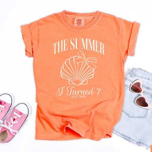 K&ouml;nnte beinhalten: Melonfarbenes T-Shirt mit dem Text "THE SUMMER I TURNED 7 EST 2019" und einer Muschel-Grafik. Das Shirt wird mit hellblauen Jeansshorts, einer rosa herzf&ouml;rmigen Sonnenbrille und rosa Turnschuhen kombiniert.