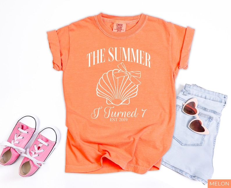 K&ouml;nnte beinhalten: Melonfarbenes T-Shirt mit dem Text "THE SUMMER I TURNED 7 EST 2019" und einer Muschel-Grafik. Das Shirt wird mit hellblauen Jeansshorts, einer rosa herzf&ouml;rmigen Sonnenbrille und rosa Turnschuhen kombiniert.