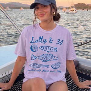 May include: A pale purple t-shirt featuring the words "Salty & 30" and blue illustrations of fish and a lemon slice. The shirt also has the text "packed with flavour since 1996".