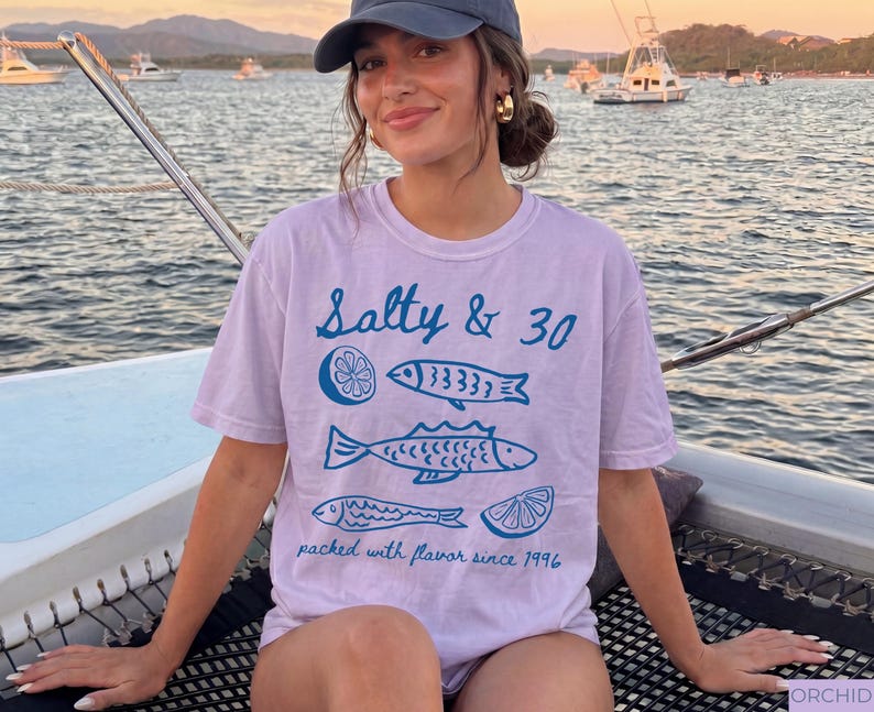 May include: A pale purple t-shirt featuring the words "Salty & 30" and blue illustrations of fish and a lemon slice. The shirt also has the text "packed with flavour since 1996".